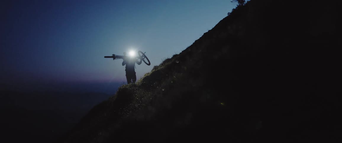 A man wearing a headlamp carries a bicycle up a hill in the dark