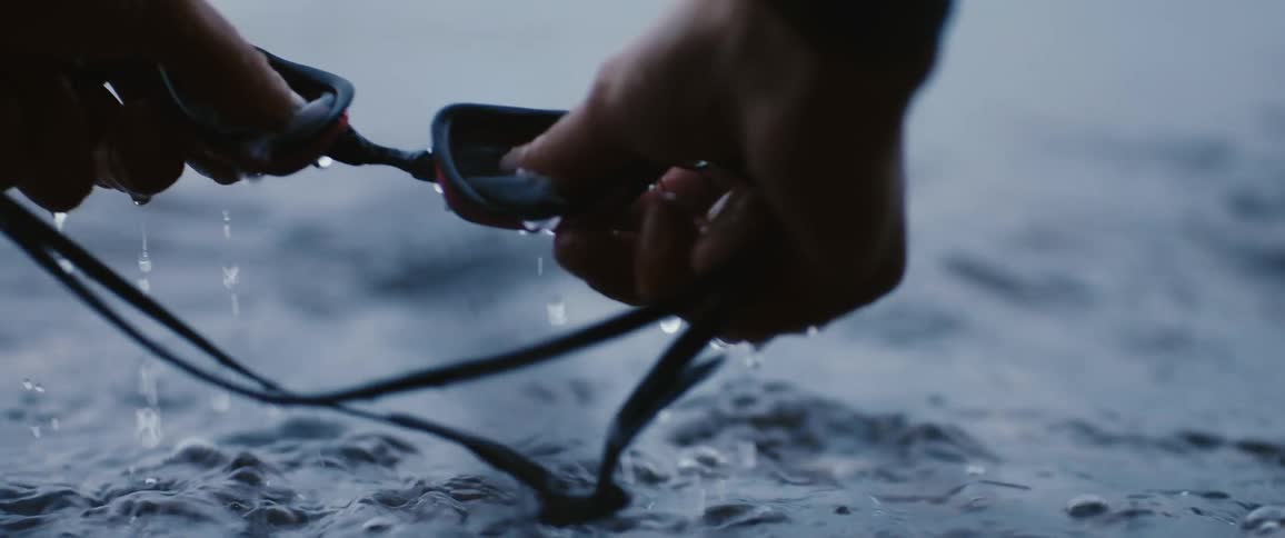 A close up of a persons hands submerging swimming goggles into wavy waters and then rubbings the lens