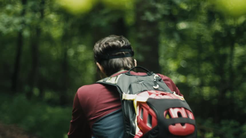 Man wearing backpack pushes bicycle along trail through woods
