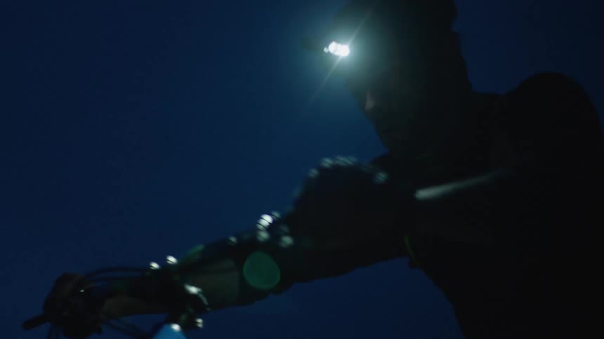 Silhouette of a man with a head lamp pushes a mountain bike uphill at night