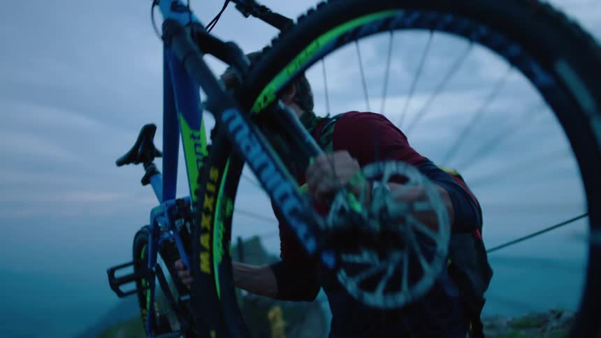 Man with blue red sweater orange shorts lifts up mountain bike on mountain road