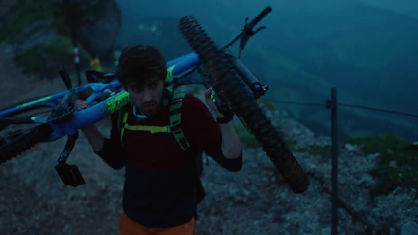 Man with bag sweater and orange pants lifts up blue bicycle on mountain