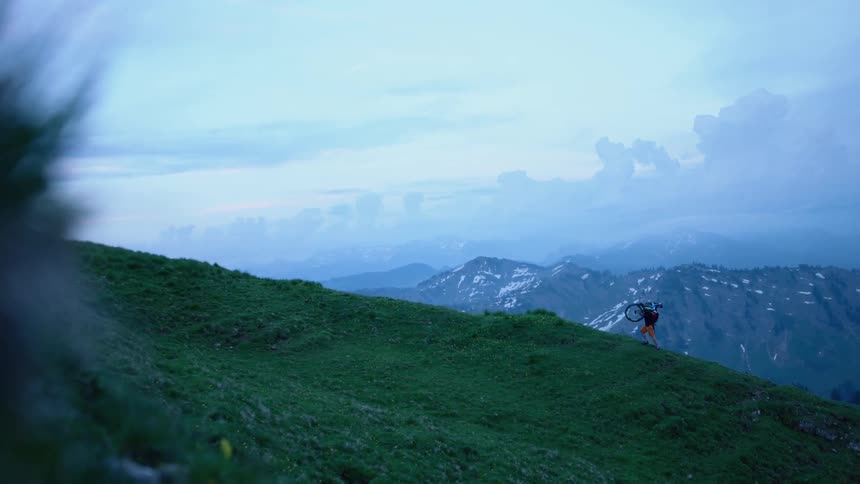Man hiking up hillside carrying mountain bike over shoulders with snowing mountains in the background and grassy terrain