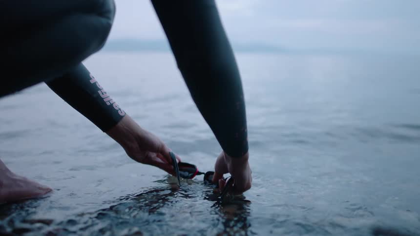 A person crouching on the shore washes their swimming goggles in a large body of water