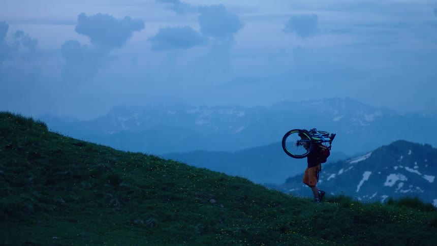 Man with backpack coat and orange shorts carries bicycle over his shoulders up mountain