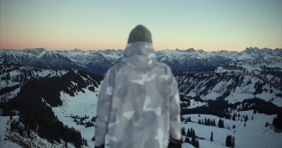 Man with wooly hat and gray coat watches snowy mountain range at dusk