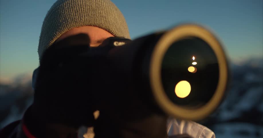 Hiker with hat and white coat takes photos with camera on snowy mountain at sunset