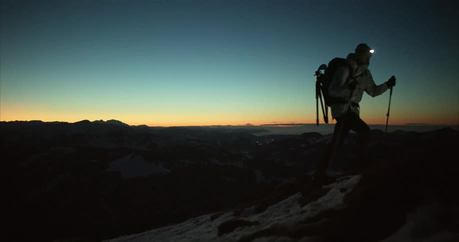 Man trekking up mountain reaches peak as the sun goes below the horizon