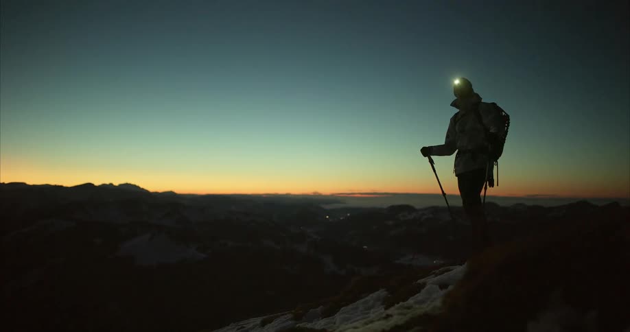 A silhouette of a hiker with ski poles a backpack and headlamp on standing looking out over mountains
