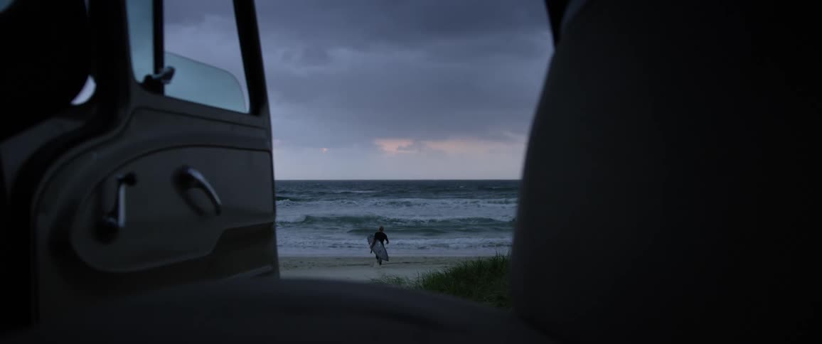 A car door open and a person holding a surf board runs on a beach toward the ocean