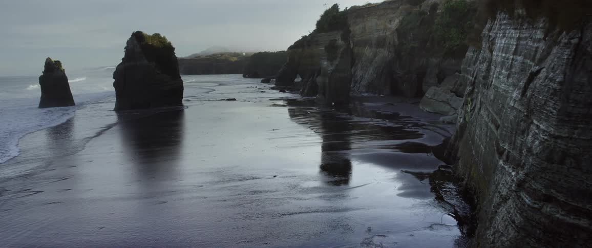 Seagulls fly among rock formations near a cliff along the coast as waves roll in to shore
