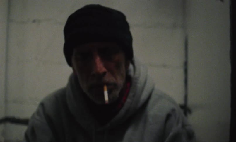 An older man wearing a black cap smoking a cigarette and rocking back and forth