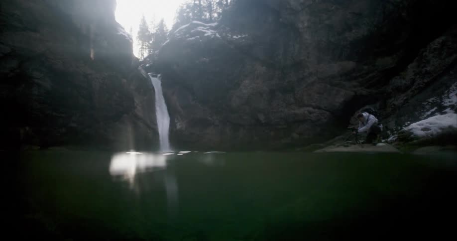 Waterfalls flow over the rocks as a man kneels near the water and the sun shines brightly in the sky