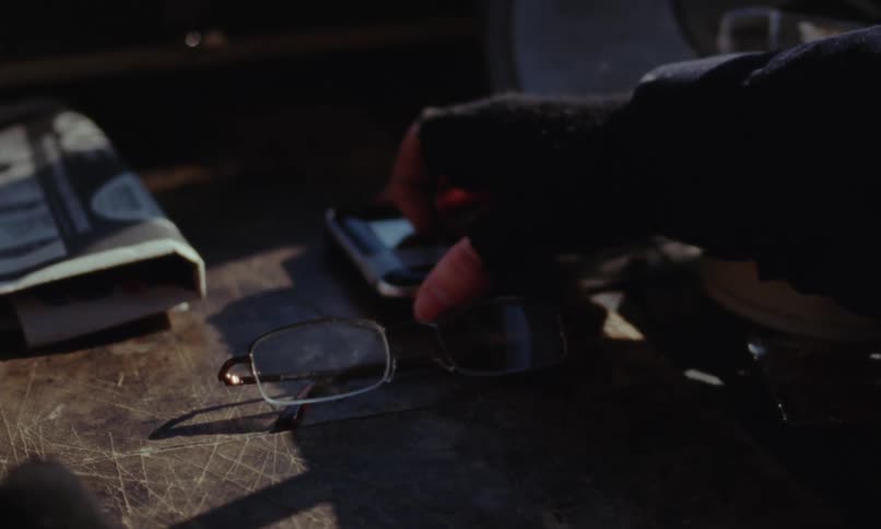 Mans hand wearing fingerless glove arranging things on a side table including a flip phone and reading glasses