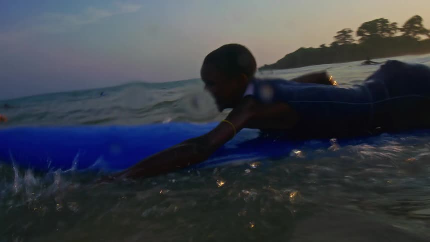 Boy with t shirt and shorts swims on blue surfboard at sunset