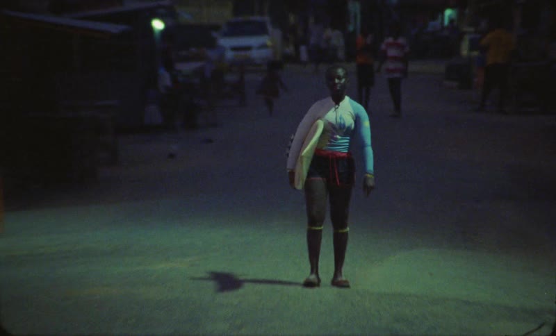 Pedestrians move along town as woman stands in street at night holding a surfboard