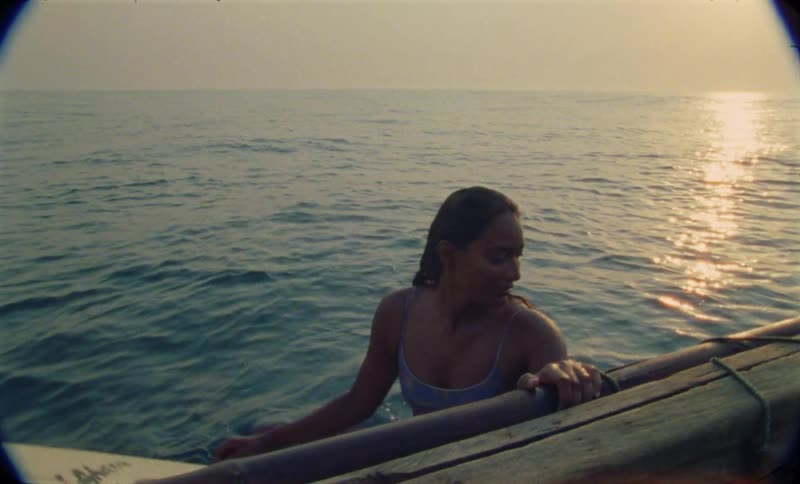 Young woman sitting over a surfboard next to a boat caught on vintage film