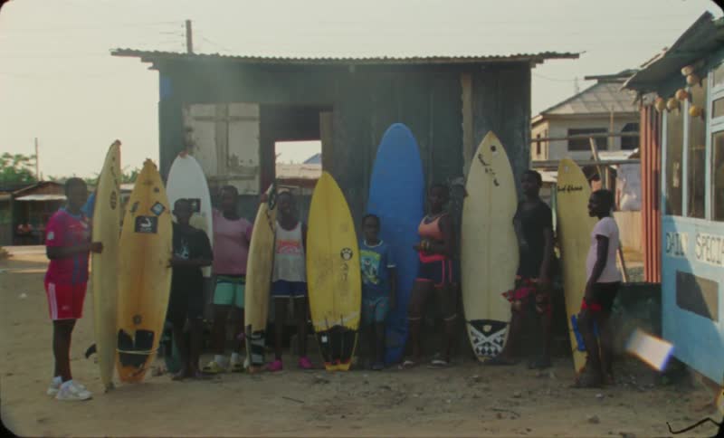 A group of kids and teenagers standing together holding up surf boards in front of a hut