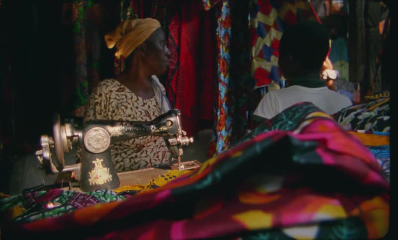 A woman behind a sewing machine talks to a boy handling clothing fabric inside a small workshop store