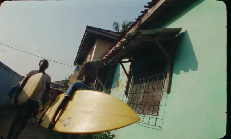 Boys walking along side walk in front of houses carrying surfboards
