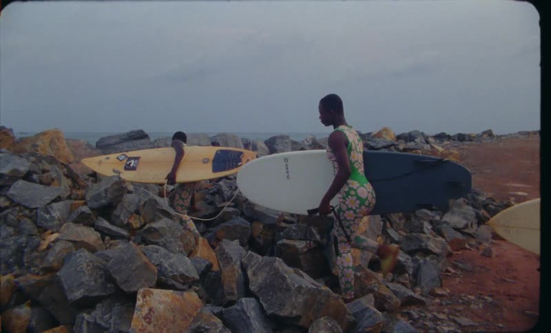 Three women carry surfboards as they climb over a mountain of large rocks on the beach