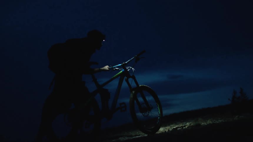 Silhouette of a man with a head lamp pushes a mountain bike uphill at night