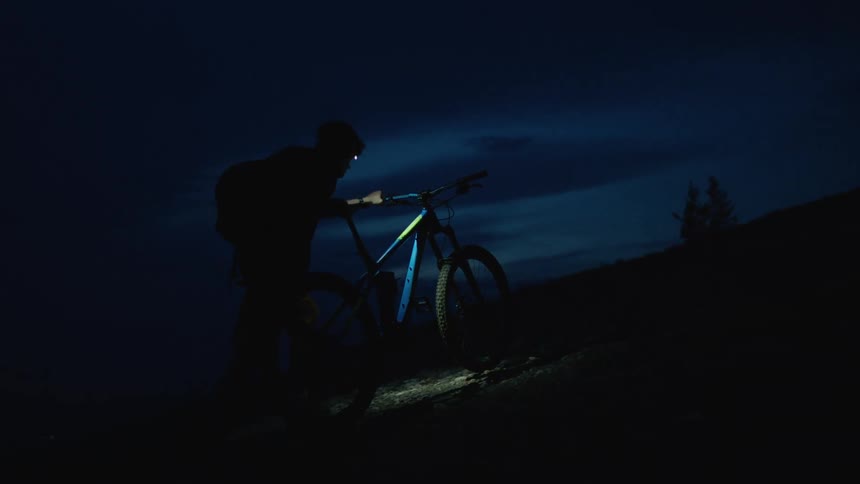 Silhouette of a man with a head lamp pushes a mountain bike uphill at night