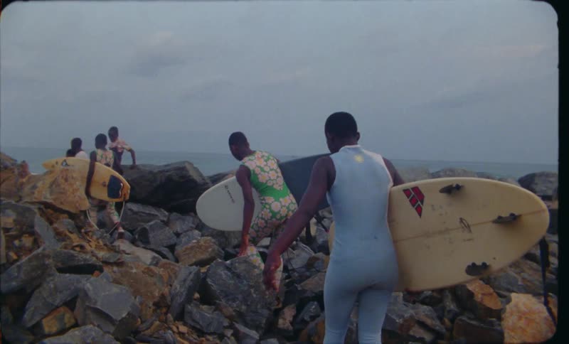 Surfers climb over rough terrain while carrying surfboards on way to shore