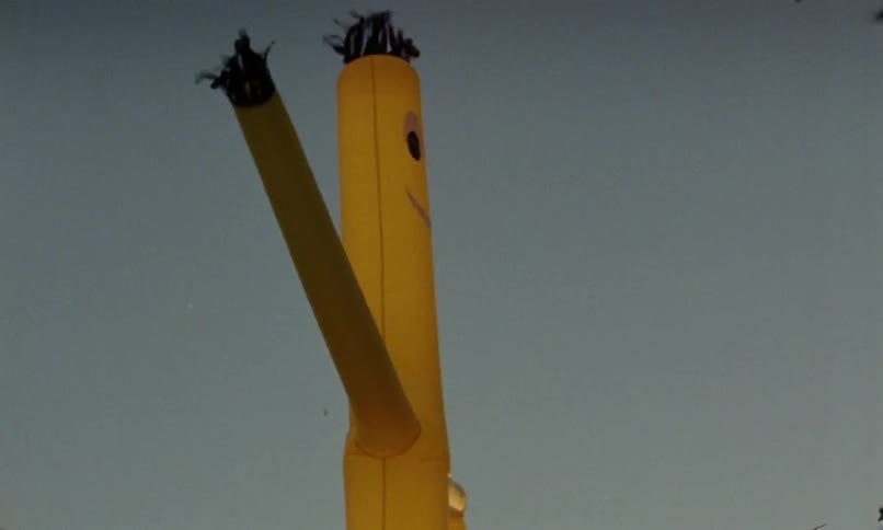 Birds fly through air as yellow tube man sways against blue sky