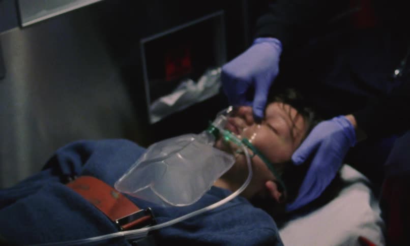 A boy is lying on a gurney in an ambulance with a breathing mask over his face while a nurse watches