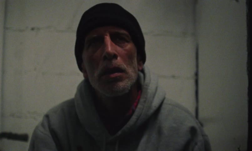 A man in an empty room smokes a cigarette as he stares straight ahead
