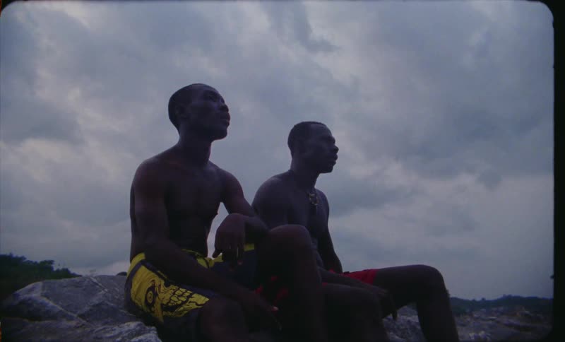 Two young men sit on a rock in their swim shorts while talking