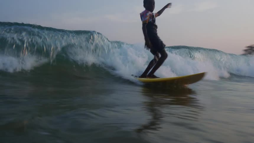A young boy on a surfboard rides a wave toward the shore at dusk