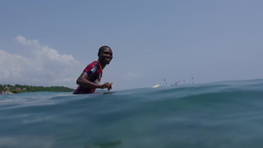 A surfer sitting on his surfboard in the ocean flashes a peace sign toward the camera