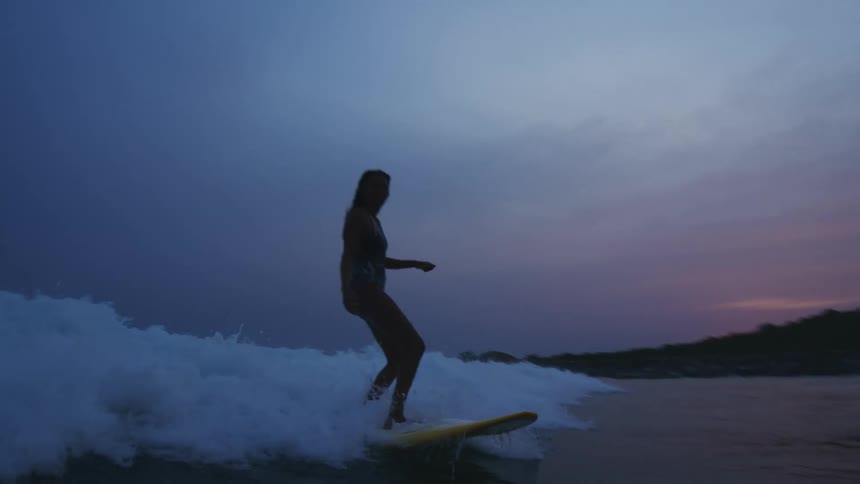 Woman rides on wave on surfboard at dusk