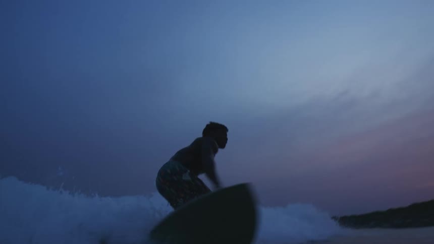 Silhouette of a young man riding a surfboard on the waves near the coast at evening