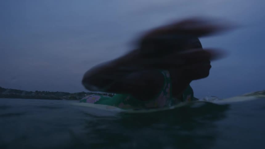 Woman wearing a colorful green wet suit swims over a surfboard at evening