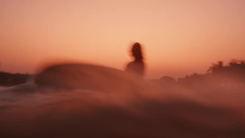 Female surfer heads out as other people surf toward beach under a red sky