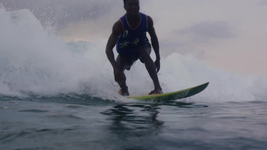 Man surfs across water ahead of wave under an overcast sky