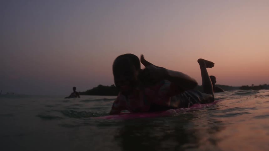 Young boy over a surfboard gives a thumb up to the camera at sunset