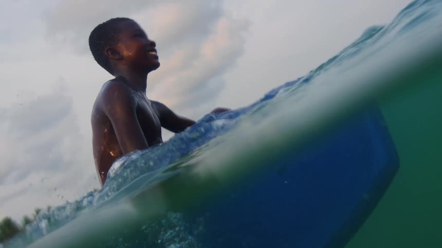 A young boy on a surfboard in the ocean during the day smiles and cheers