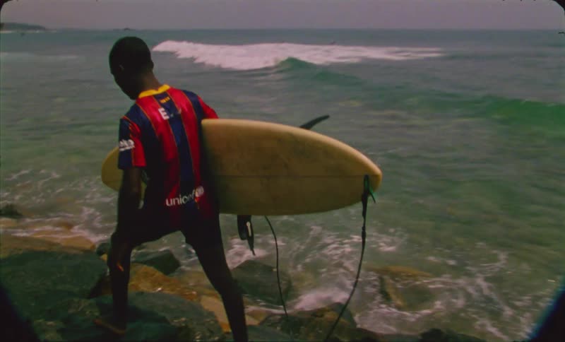 A teenage boy is carrying a surfboard as he walks on rocks to the water along the beach