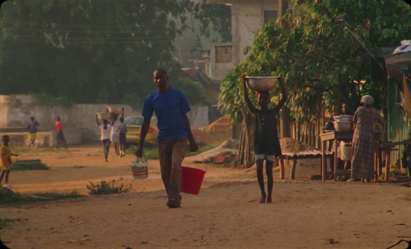 A boy and his father carry buckets of water and baskets through a dirtlined thirdworld town
