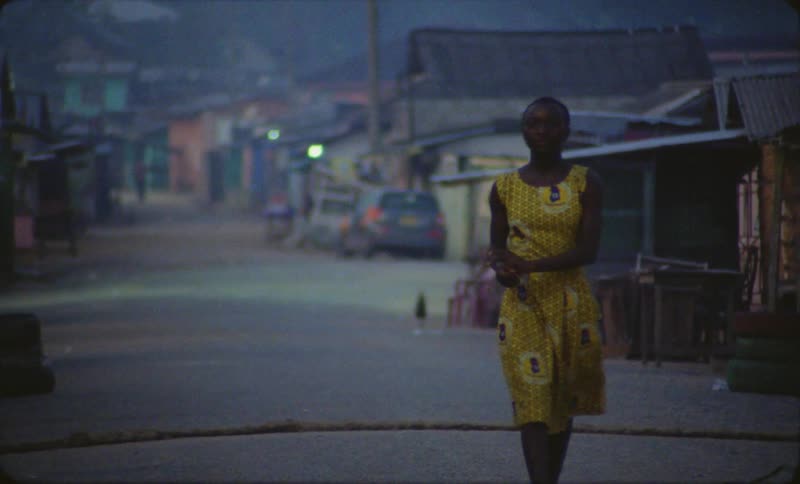 A woman in a yellow dress walks down the street of her neighborhood in a third world nation