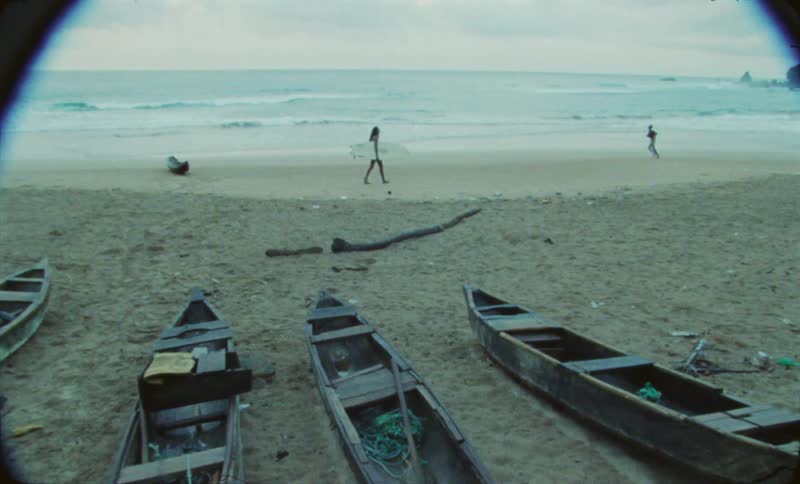 Four wooden boats parked in the sand on the beach as a woman carryinga surfboard walks across the sand as a child runs around in front of her