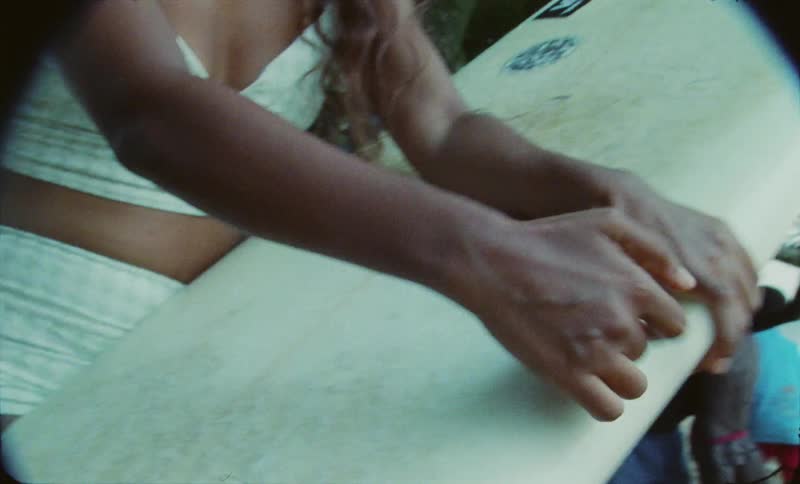 A woman is waxing her surfboard at the beach as children play near a tree