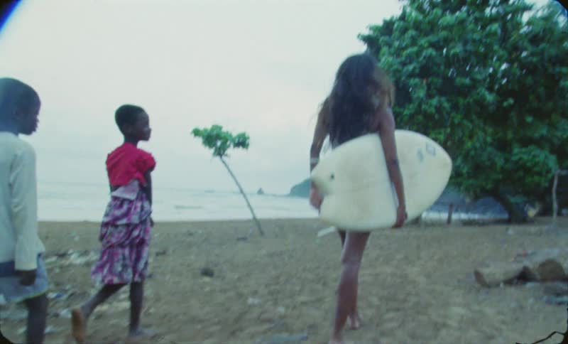 A teenage girl is carrying her surfboard to the beach as two young children follow her