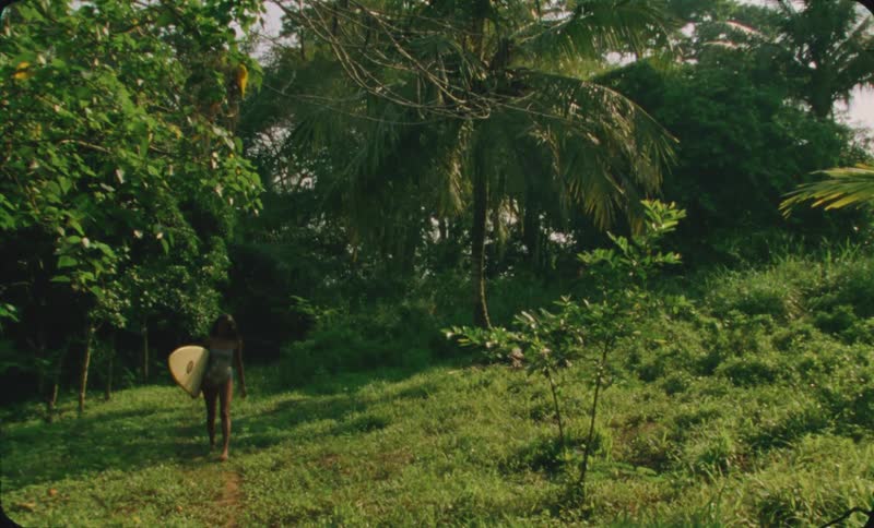 A woman carrying a surf board walking through a dense green forest