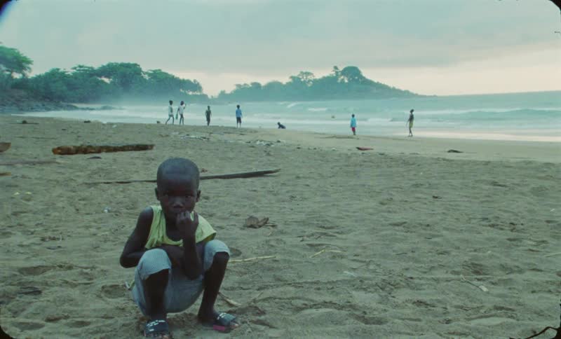 Boy crouches on beach as other kids play soccer in the background