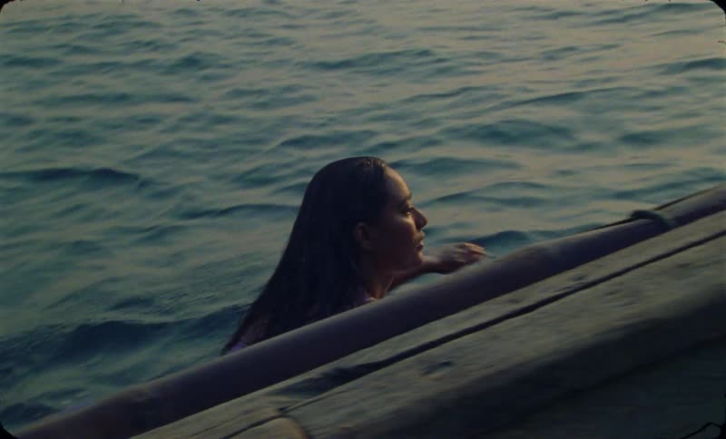 A teenage girl is lying on her surfboard as she paddles with her arms by a boat as the sun shines brightly on the horizon